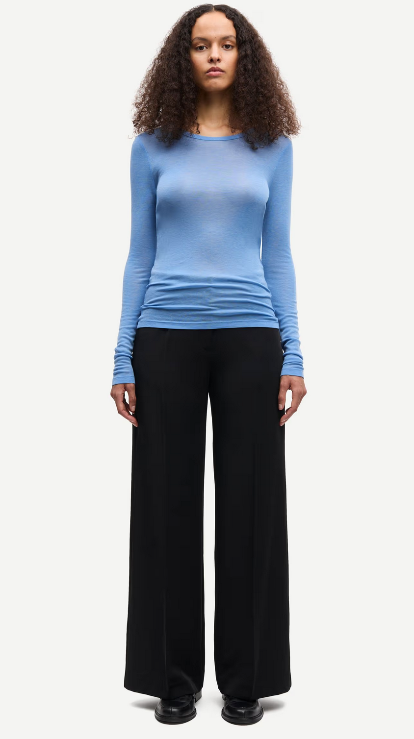 A person stands facing forward, wearing the Samsøe Samsøe Sadou T-shirt Long Sleeve in Della Robbia Blue, paired with wide-leg black pants and black shoes against a plain light background.