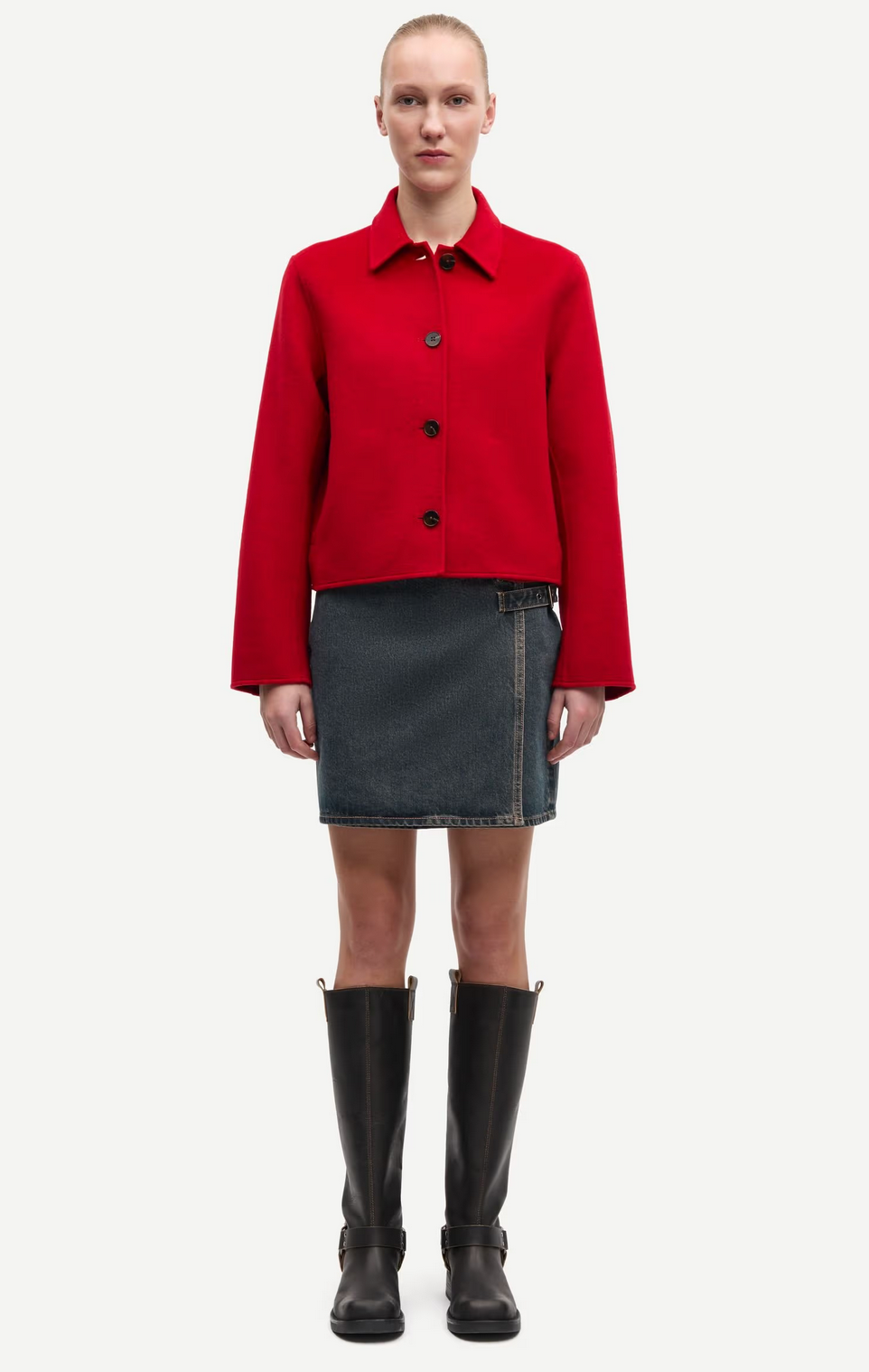 A person stands facing forward against a plain white background, wearing the Saholly Jacket in Haute Red by Samsøe Samsøe, paired with a dark denim mini skirt and tall black boots.