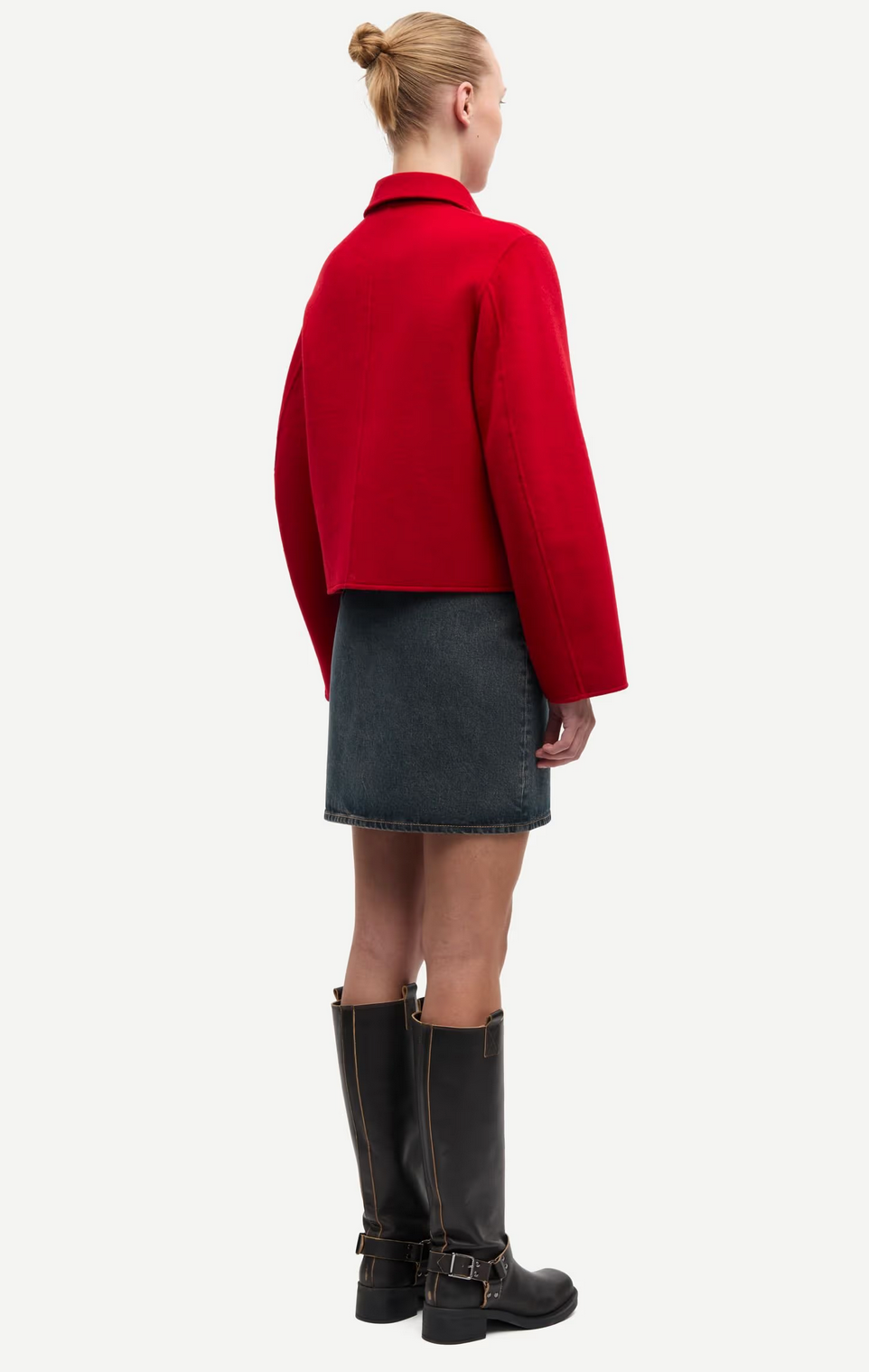 A person stands facing away, wearing the Samsøe Samsøe Saholly Jacket in Haute Red, paired with a dark denim skirt and black knee-high boots against a plain background.