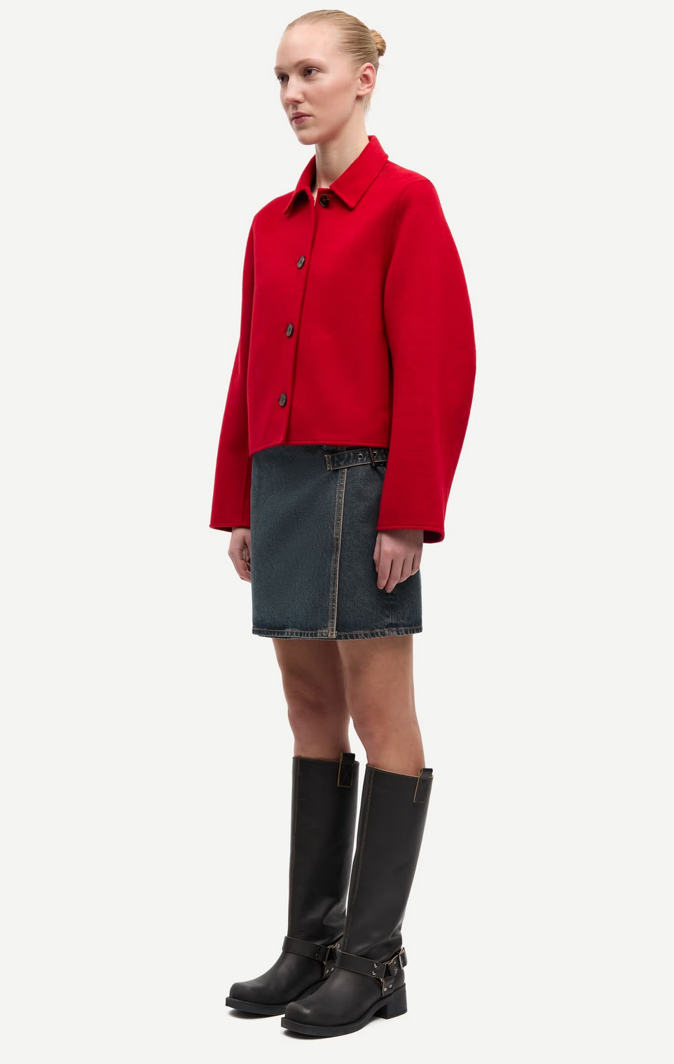 A woman stands against a plain white background wearing the Saholly Jacket - Haute Red by Samsøe Samsøe, paired with a dark denim skirt and knee-high black boots.