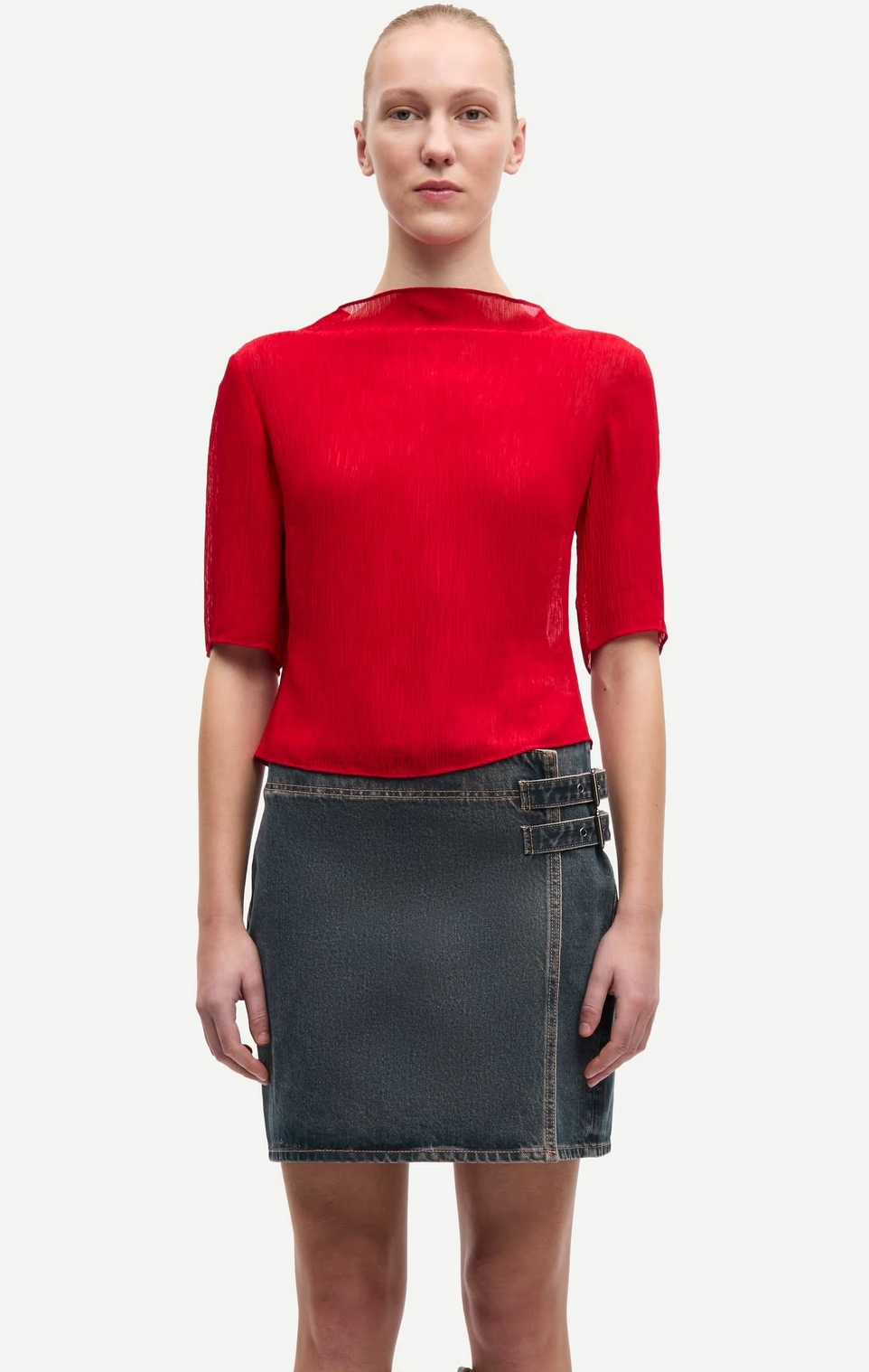A person stands facing forward, wearing the Samsøe Samsøe Samiller Top in Haute Red—a short-sleeved, semi-sheer top crafted from recycled viscose—and a dark denim mini skirt with side buckles, set against a plain white background.