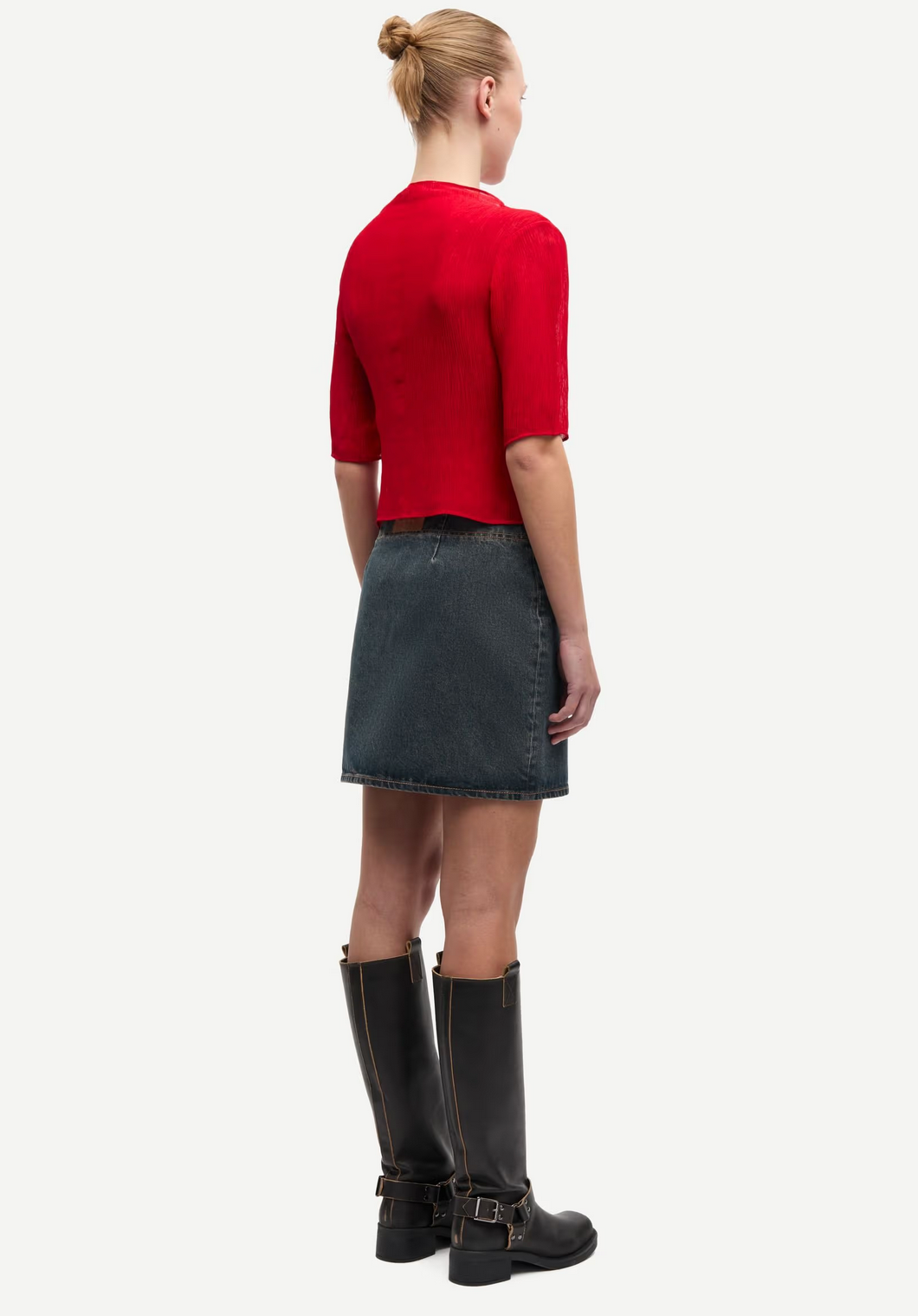 Facing away against a white background, the person wears the Samsøe Samsøe Samiller Top in Haute Red, a short-sleeved semi-sheer recycled viscose piece, with a dark denim skirt and black buckled knee-high boots; hair in a bun.