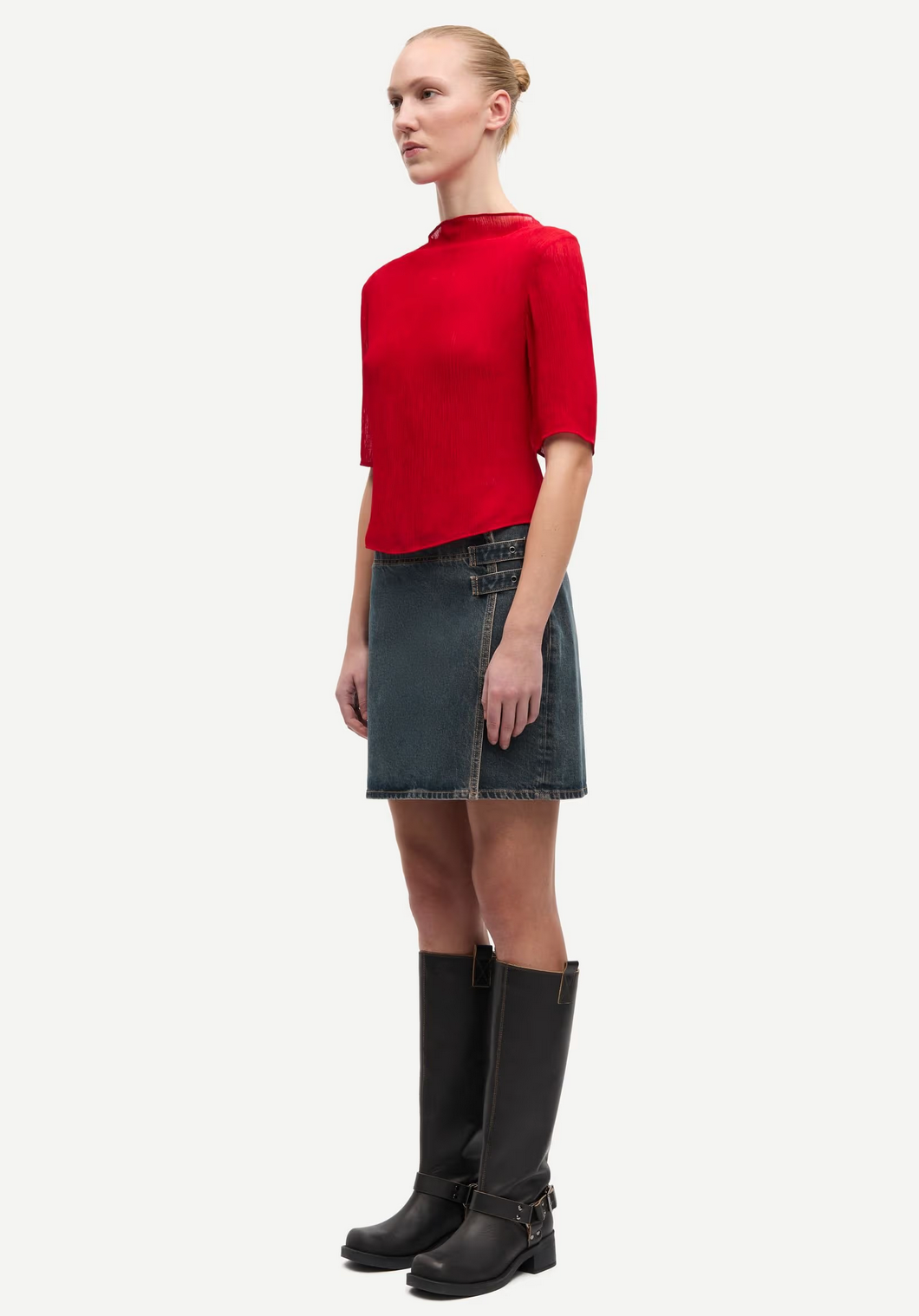 A woman stands in profile wearing the Samsøe Samsøe Samiller Top in Haute Red, a blue denim mini skirt with a front slit, and knee-high black boots with side zippers.