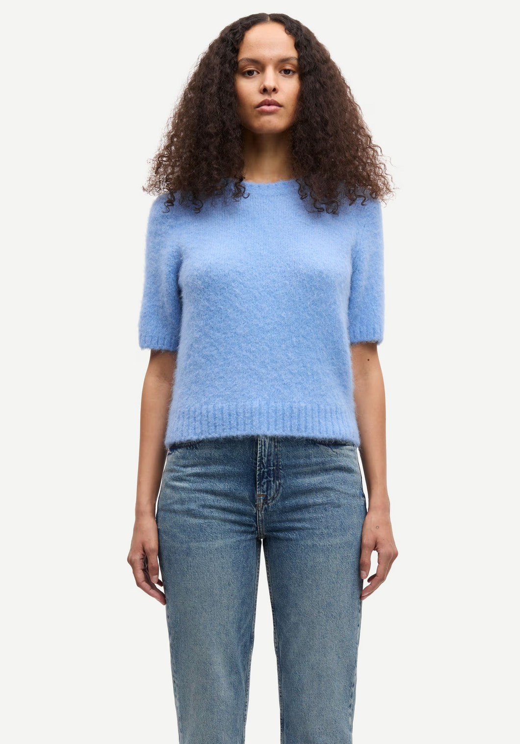 A person with curly hair wears the Samsøe Samsøe Sajeanne Sweater in Della Robbia Blue and blue jeans, standing against a plain white background.