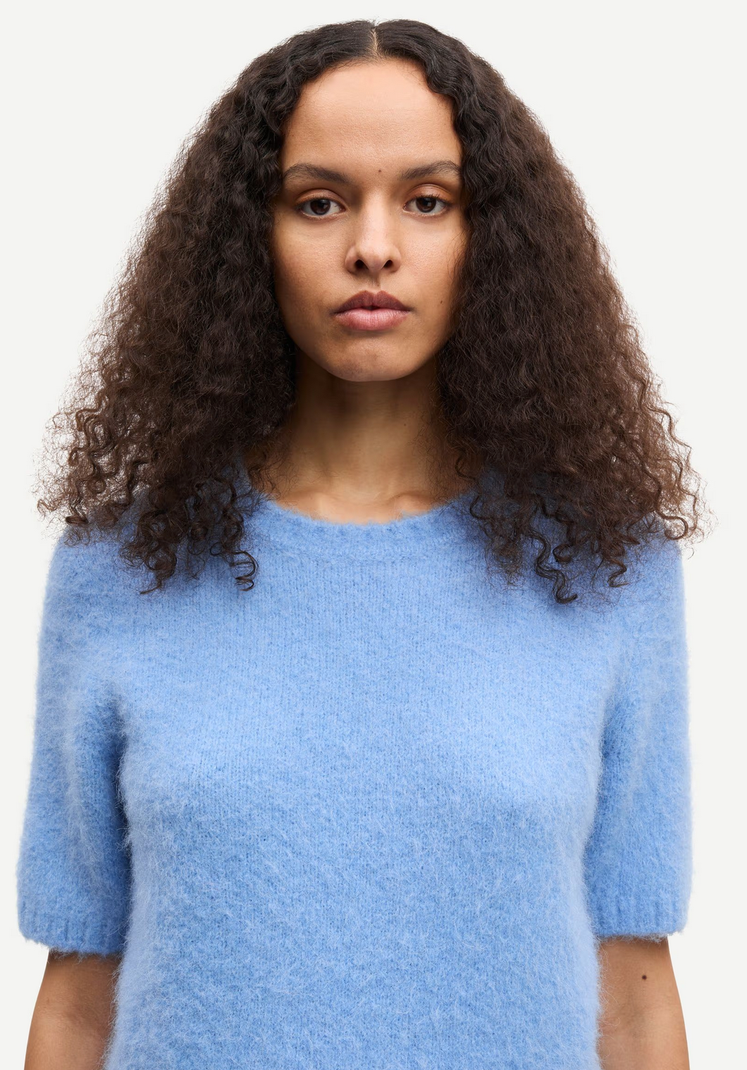 A person with long curly hair wears the Samsøe Samsøe Sajeanne Sweater in Della Robbia Blue, facing forward against a plain background.