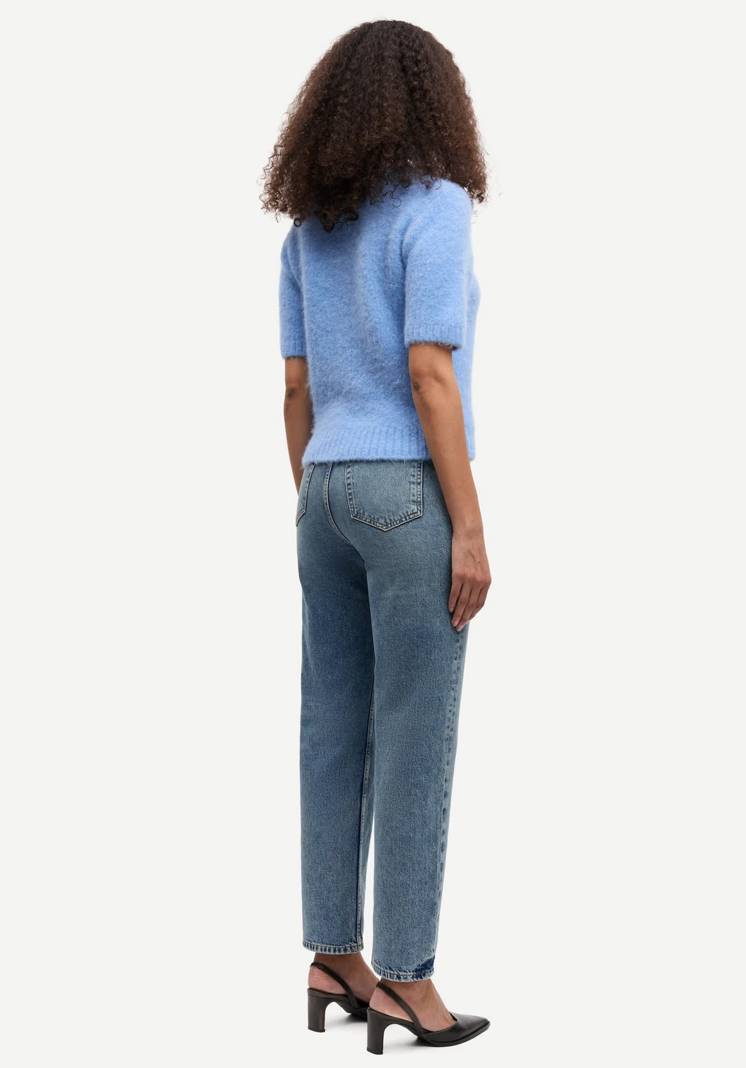 A person with curly hair, wearing the Samsøe Samsøe Sajeanne Sweater in Della Robbia Blue, blue jeans, and black heels, stands facing away from the camera against a plain white background.