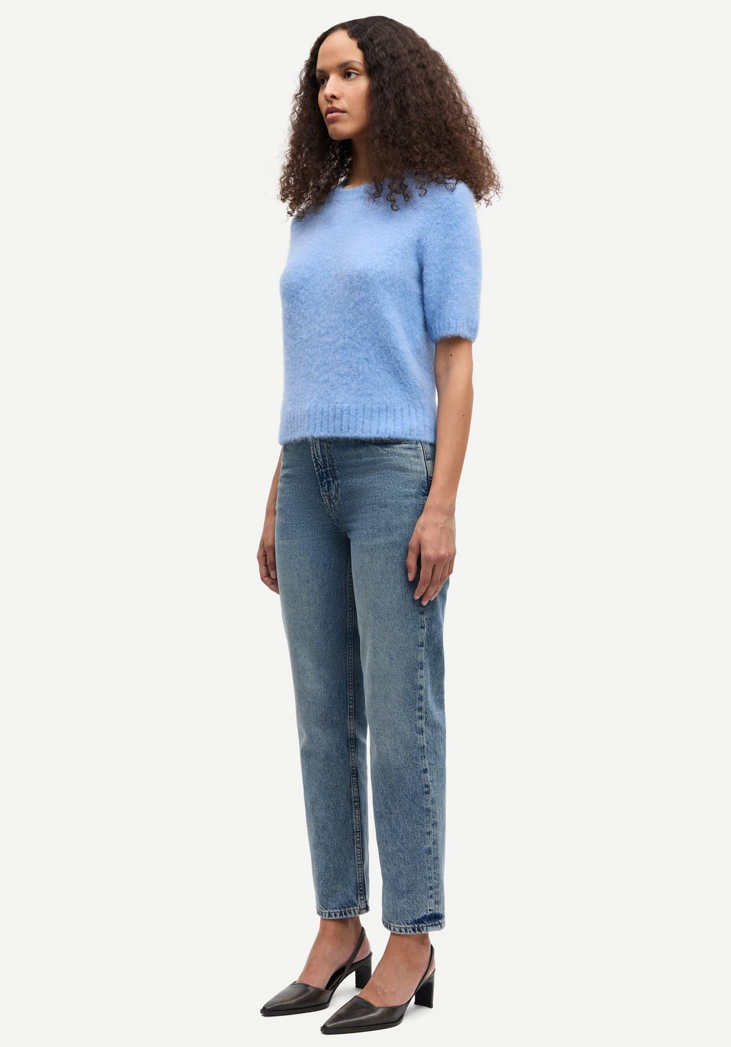 A woman stands against a white background wearing the Samsøe Samsøe Sajeanne Sweater in Della Robbia Blue, paired with blue jeans and black heels.