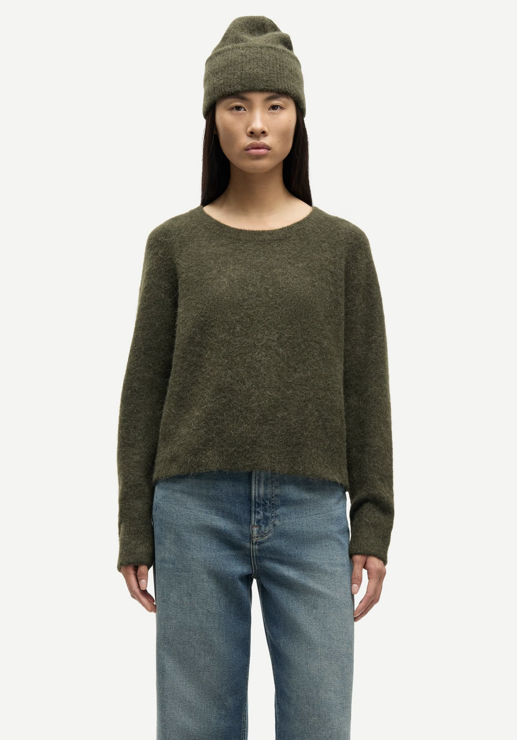 A person stands facing forward against a plain background, wearing a Samsøe Samsøe Nor - O-Neck Short Sweater in Deep Forest, a matching beanie, and blue jeans.