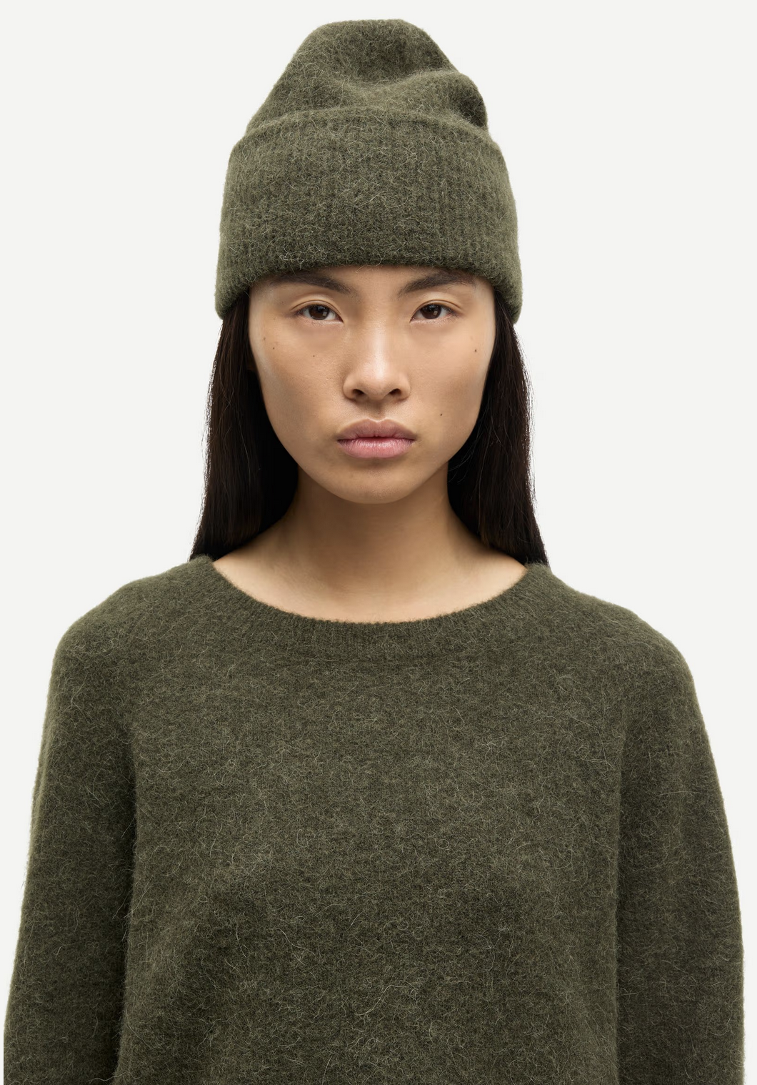 A person with long dark hair wears the Samsøe Samsøe Nor - O-Neck Short Sweater in Deep Forest and a matching beanie, facing forward with a neutral expression against a plain background.