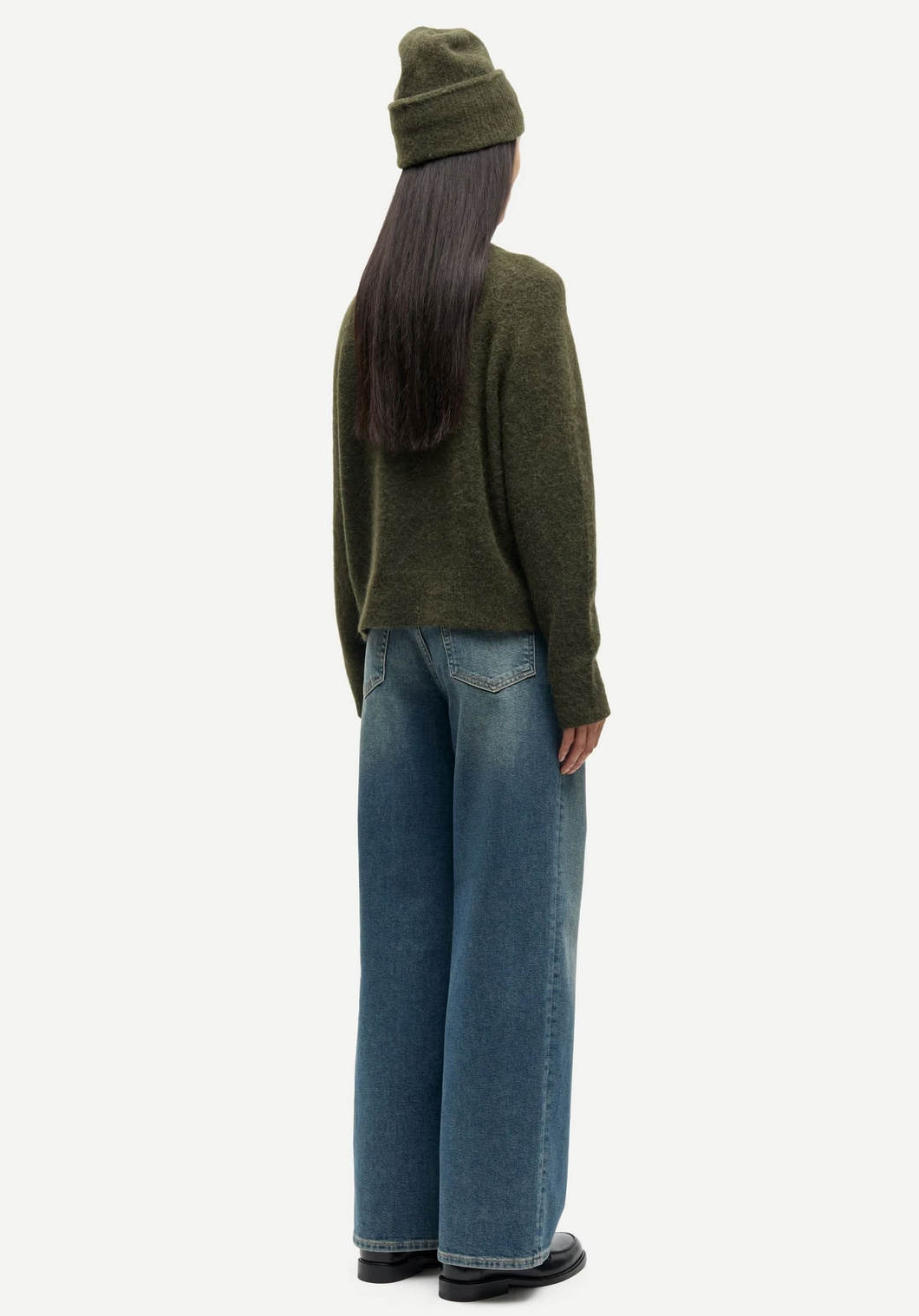 A person stands facing away against a plain background, wearing an olive green beanie, Samsøe Samsøe's Nor - O-Neck Short Sweater in Deep Forest, wide-leg blue jeans, and black shoes.