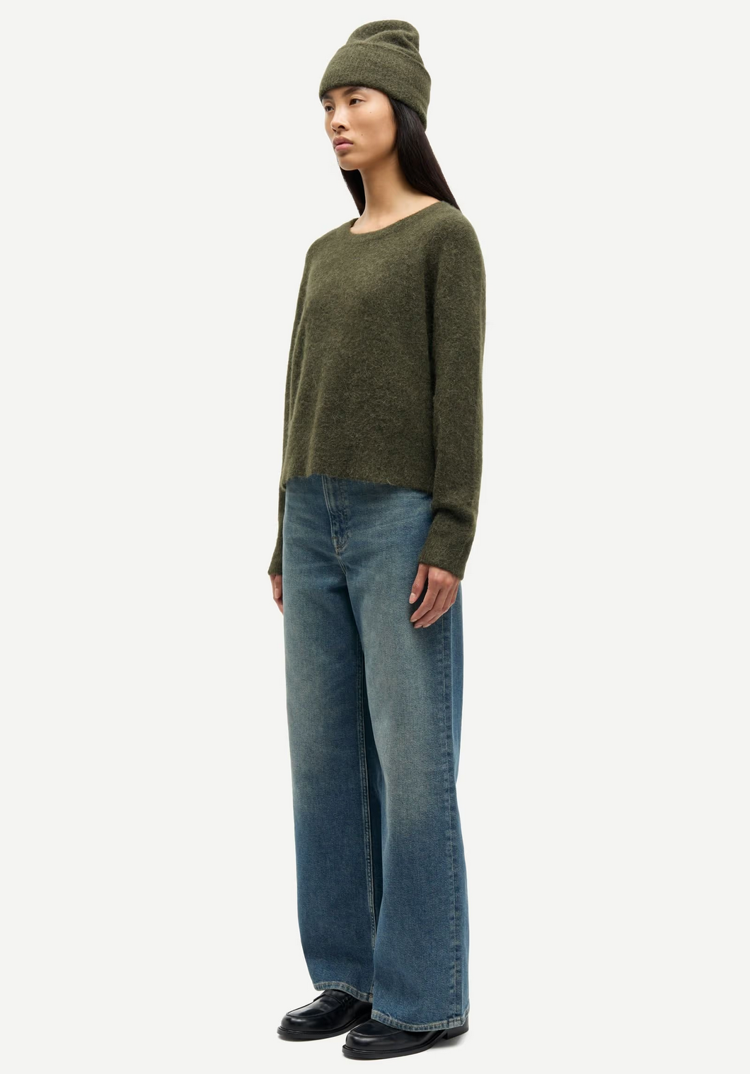 A woman wears a Samsøe Samsøe Nor-O-Neck Short Sweater in Deep Forest, paired with a green beanie, wide-leg blue jeans, and black shoes, standing against a plain white background.