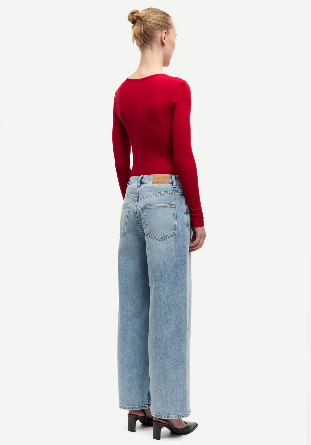 A person with hair in a bun wears the Sadou T-shirt Long Sleeve in Haute Red by Samsøe Samsøe, paired with light blue wide-leg TENCEL Lyocell jeans and black-heeled shoes, standing against a plain white background.