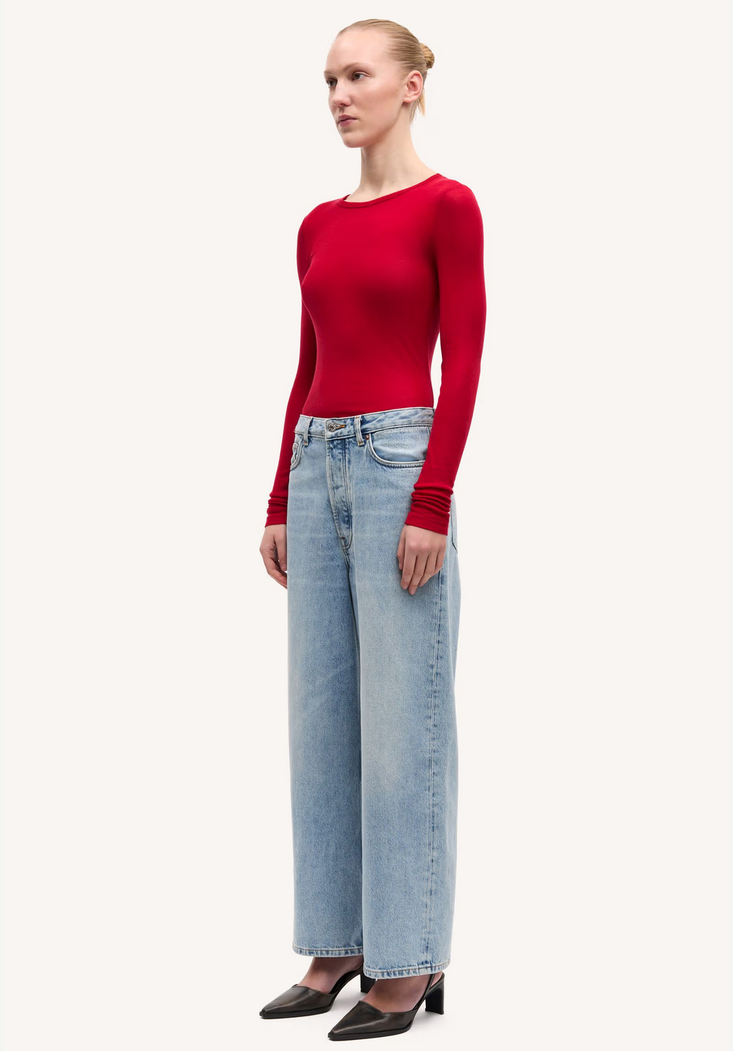 A person stands slightly sideways, wearing the Samsøe Samsøe Sadou T-shirt Long Sleeve in Haute Red, paired with light blue wide-leg jeans and black heeled shoes against a plain background.