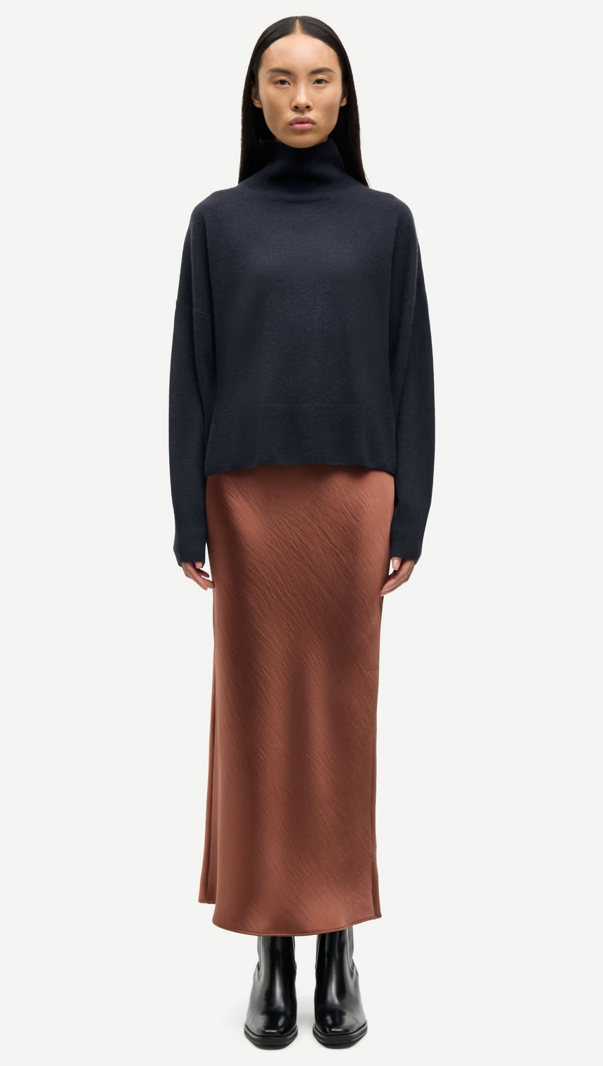 A woman stands facing forward wearing a navy turtleneck sweater, Samsøe Samsøe’s Saagneta Long Satin Skirt in Tortoise Shell, and black ankle boots against a plain white background.