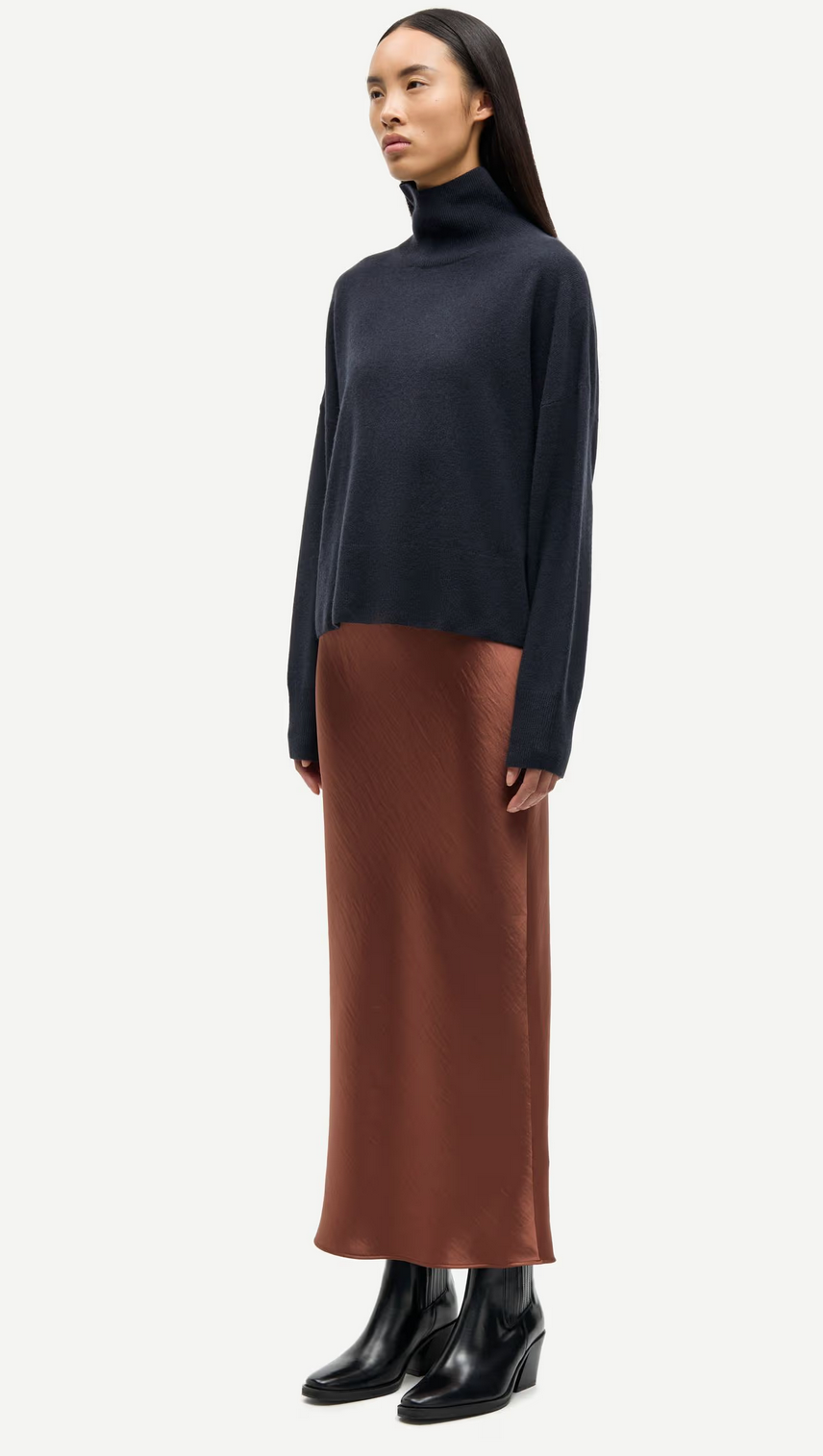A woman wears a dark turtleneck, black ankle boots, and the Samsøe Samsøe Saagneta Long Satin Skirt in Tortoise Shell, standing against a plain background.