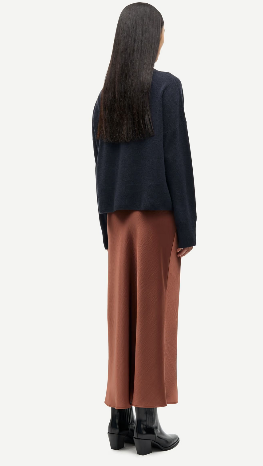 A person with long dark hair wears a loose dark sweater, black boots, and the Saagneta Long Satin Skirt in Tortoise Shell by Samsøe Samsøe, standing with their back to the camera.