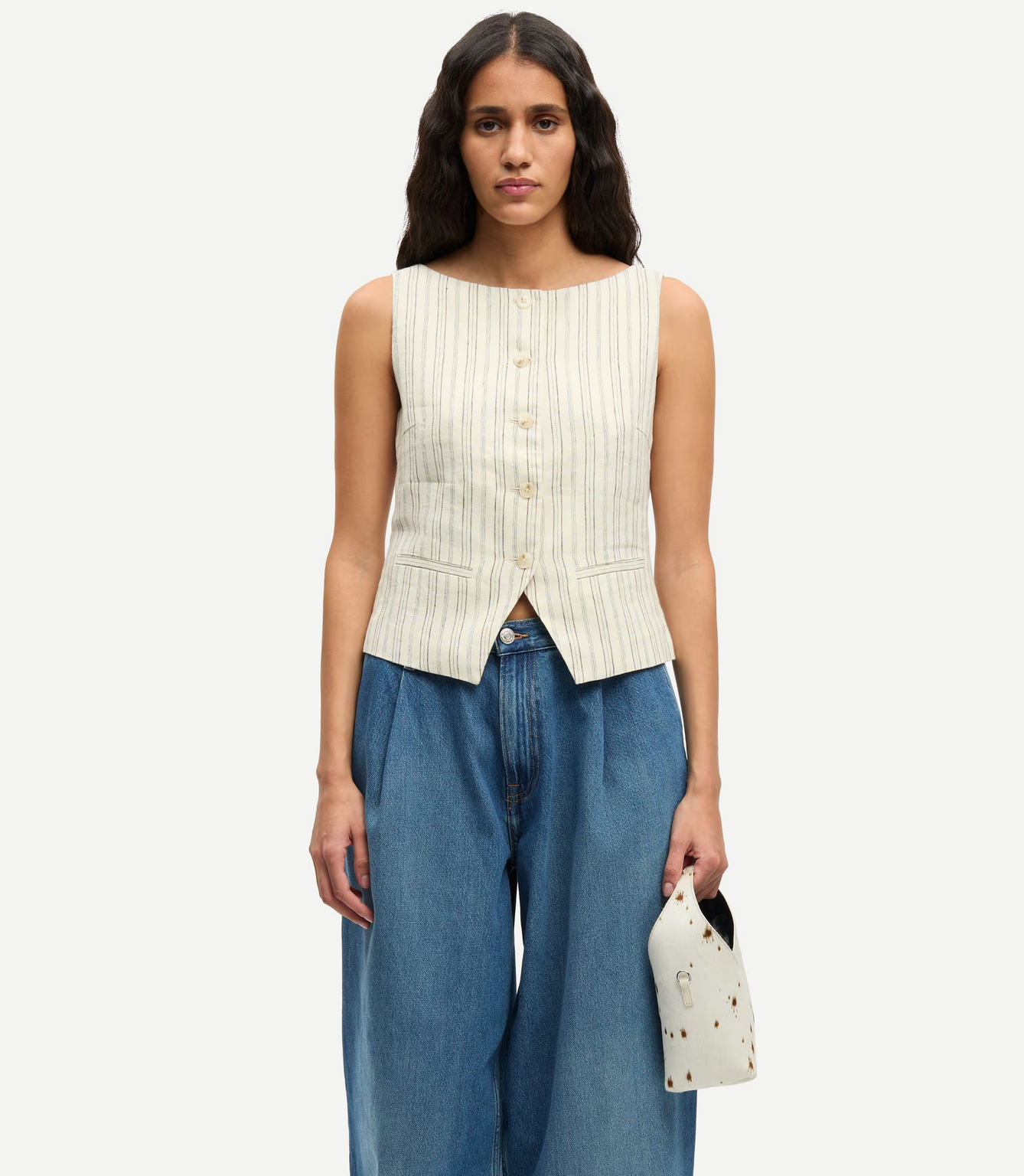 A woman wears the Saloves Vest - Bone by Samsøe Samsøe, which is sleeveless and woven with a button closure, paired with wide-leg blue jeans and holding a small white bag with brown speckles.