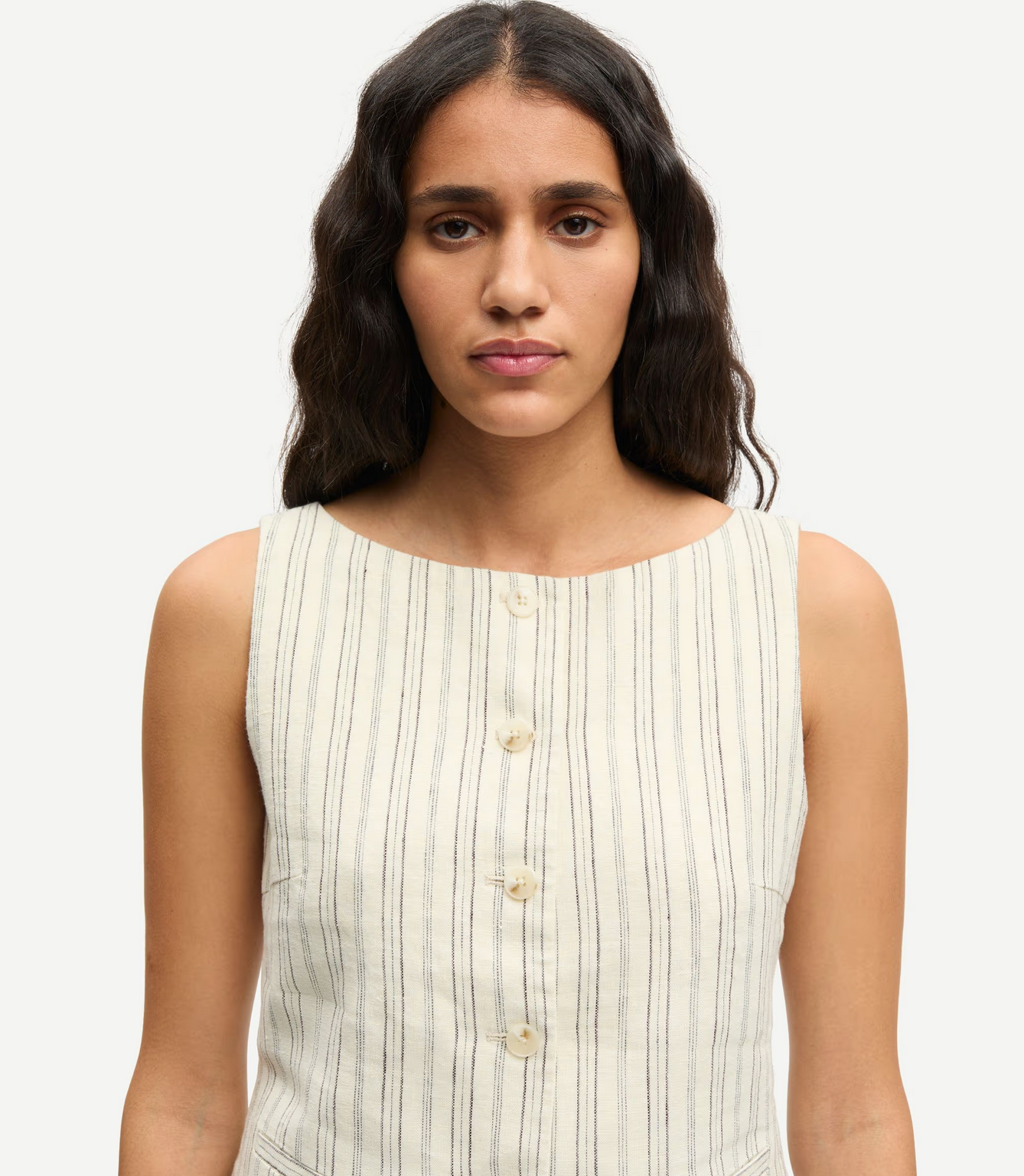 A woman with long, wavy dark hair wears the Samsøe Samsøe Saloves Vest in Bone, a sleeveless cream vest with vertical stripes and button closure, facing the camera against a plain background.