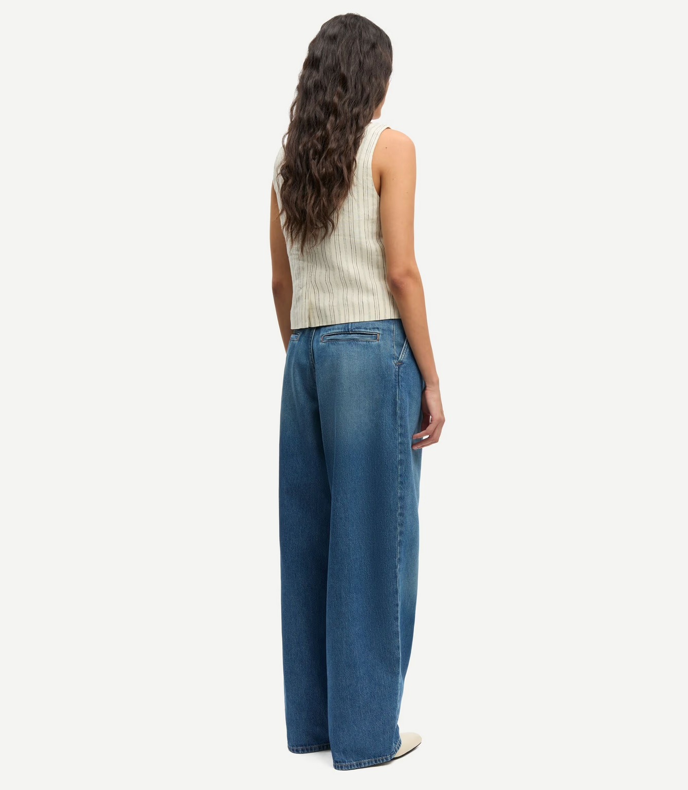 A person stands with back turned, wearing the Samsøe Samsøe Saloves Vest in Bone, paired with wide-leg blue jeans and white shoes, against a plain white background.