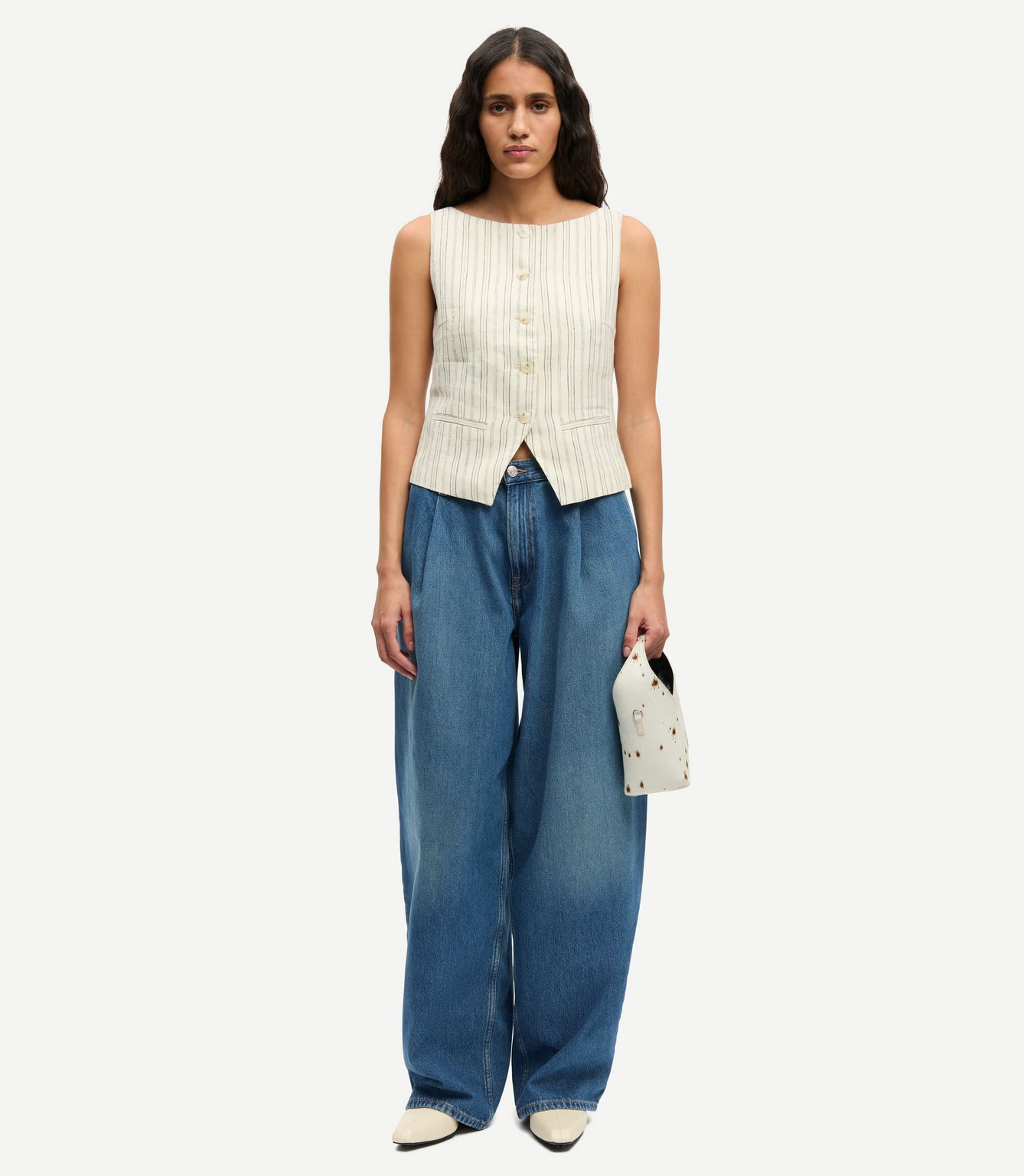A woman stands against a plain background wearing the Samsøe Samsøe Saloves Vest in Bone, styled with wide-leg blue jeans, white pointed shoes, and a small white handbag.