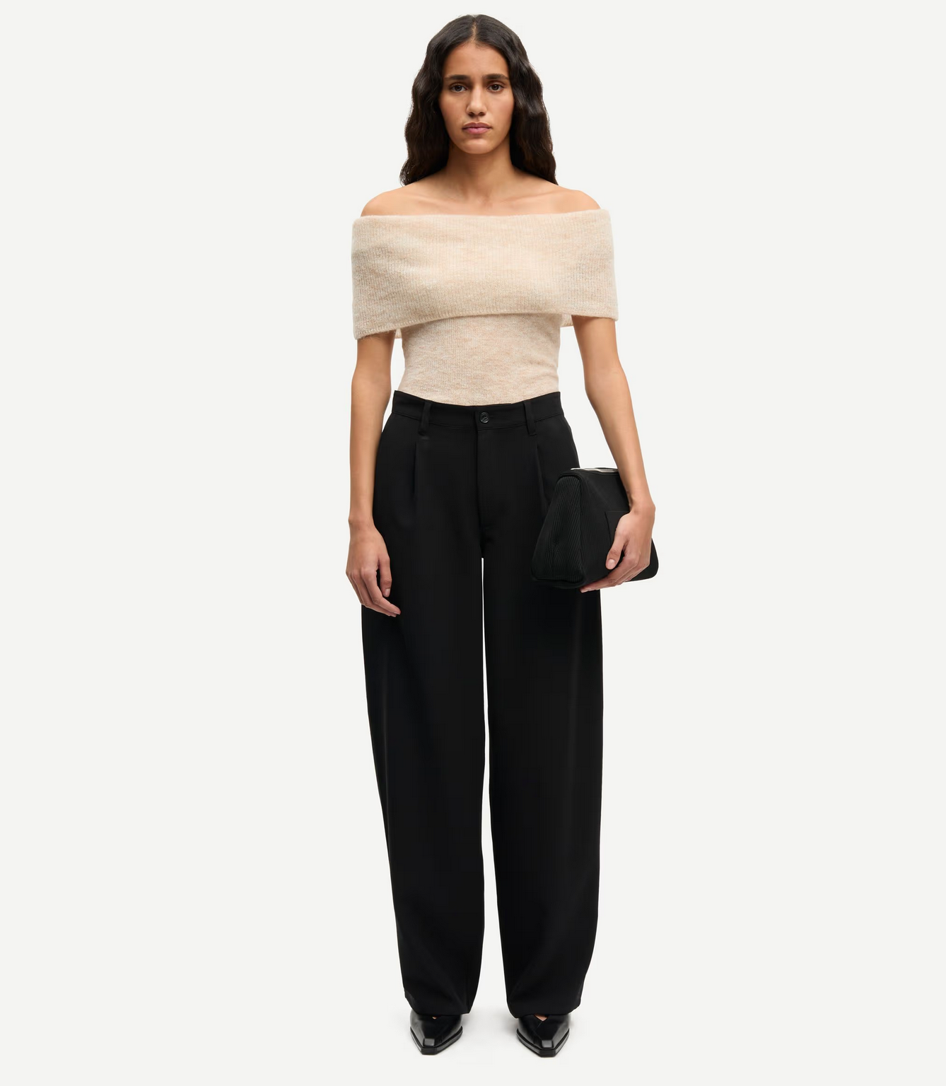 A woman stands facing forward, wearing an off-the-shoulder beige top, Saharper Wide Trousers in black by Samsøe Samsøe, black shoes, and holding a black clutch bag.