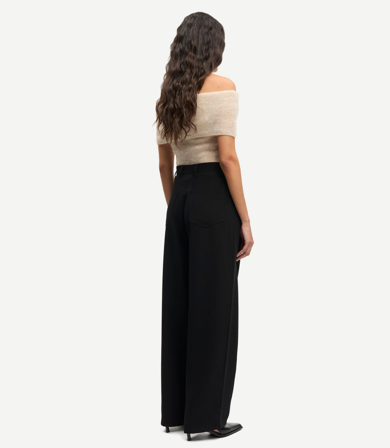 A woman with long wavy hair stands facing away, wearing an off-shoulder beige top and Samsøe Samsøe Saharper Wide Trousers in black made from recycled polyester, paired with black shoes against a plain white background.