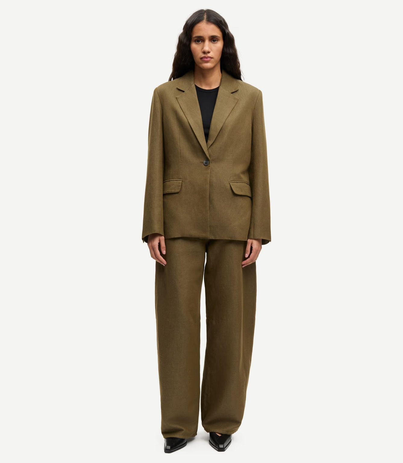 A woman faces forward wearing Samsøe Samsøe’s Saroko Trousers in Overland Trek Melange, styled with an olive green oversized single-button blazer, black top, and black shoes.