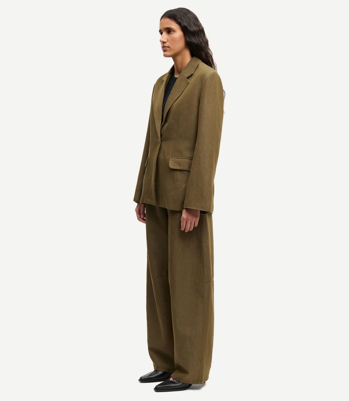 A person stands in profile wearing Samsøe Samsøe's Saroko Trousers in Overland Trek Melange, styled with an oversized olive green suit and black shoes, set against a plain white background.