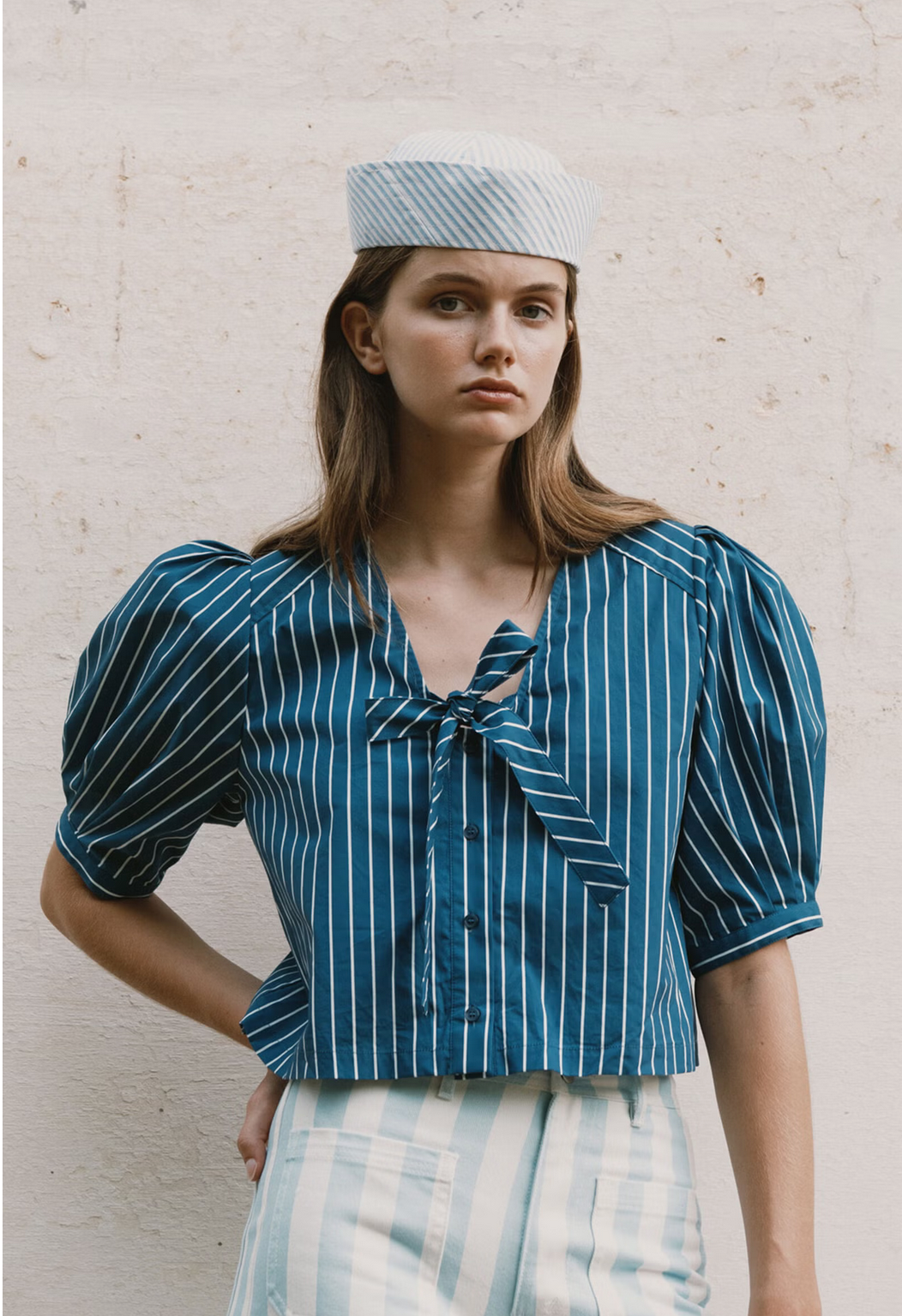 A woman wears the Tiny Big Sister Tie Detail Blouse in navy with puffed sleeves and a bow, paired with a matching skirt and a light blue hat, standing against a light wall.