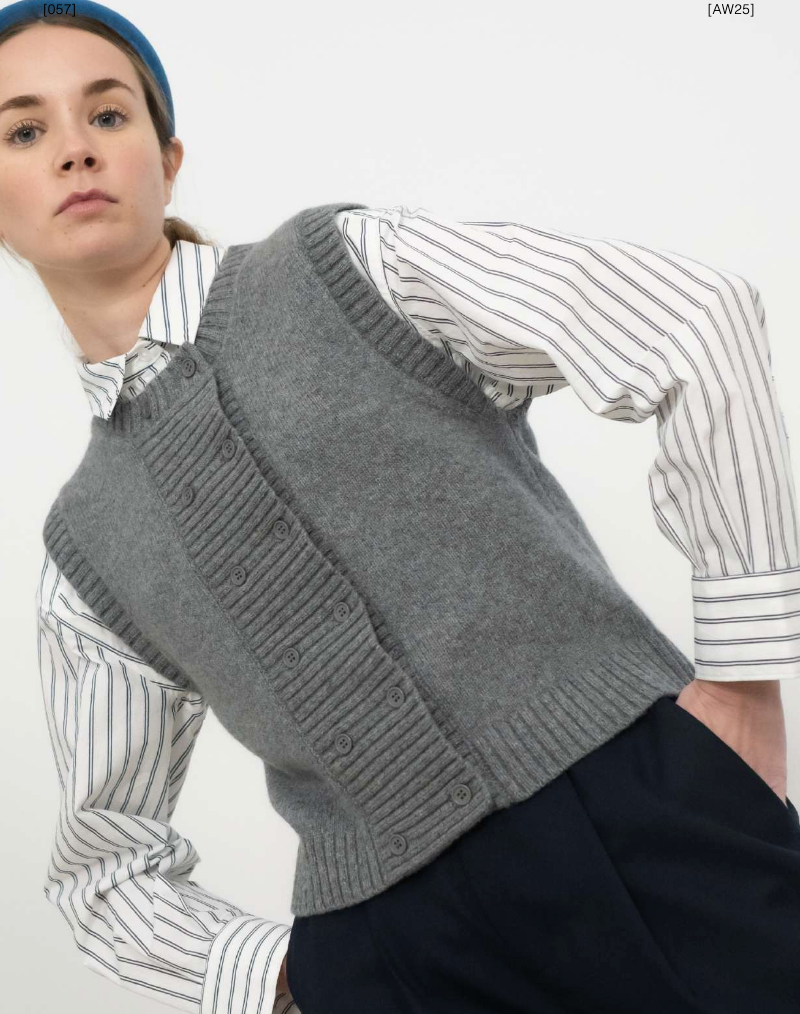 A person wears a blue headband, Tiny Big Sister Buttoned Vest in dark grey, a white and grey striped shirt, and dark trousers while posing against a plain background.