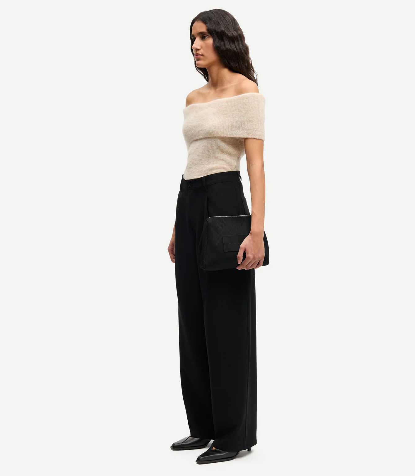 A woman stands in a neutral pose, wearing a beige off-the-shoulder top and Samsøe Samsøe’s Saharper Wide Trousers in black, paired with black pointed shoes and holding a black clutch.