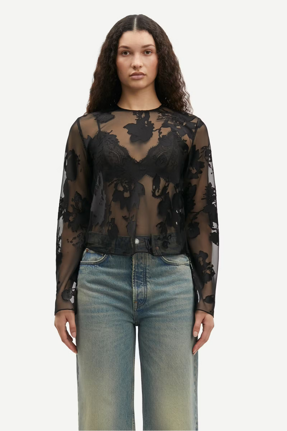 a women with brown hair wears a black sheer blouse with flower pattern and blue jeans samsoe samsoe