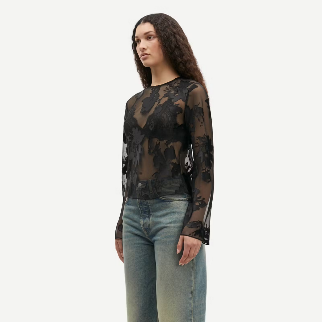 a women with brown hair wears a black sheer blouse with flower pattern and blue jeans samsoe samsoe