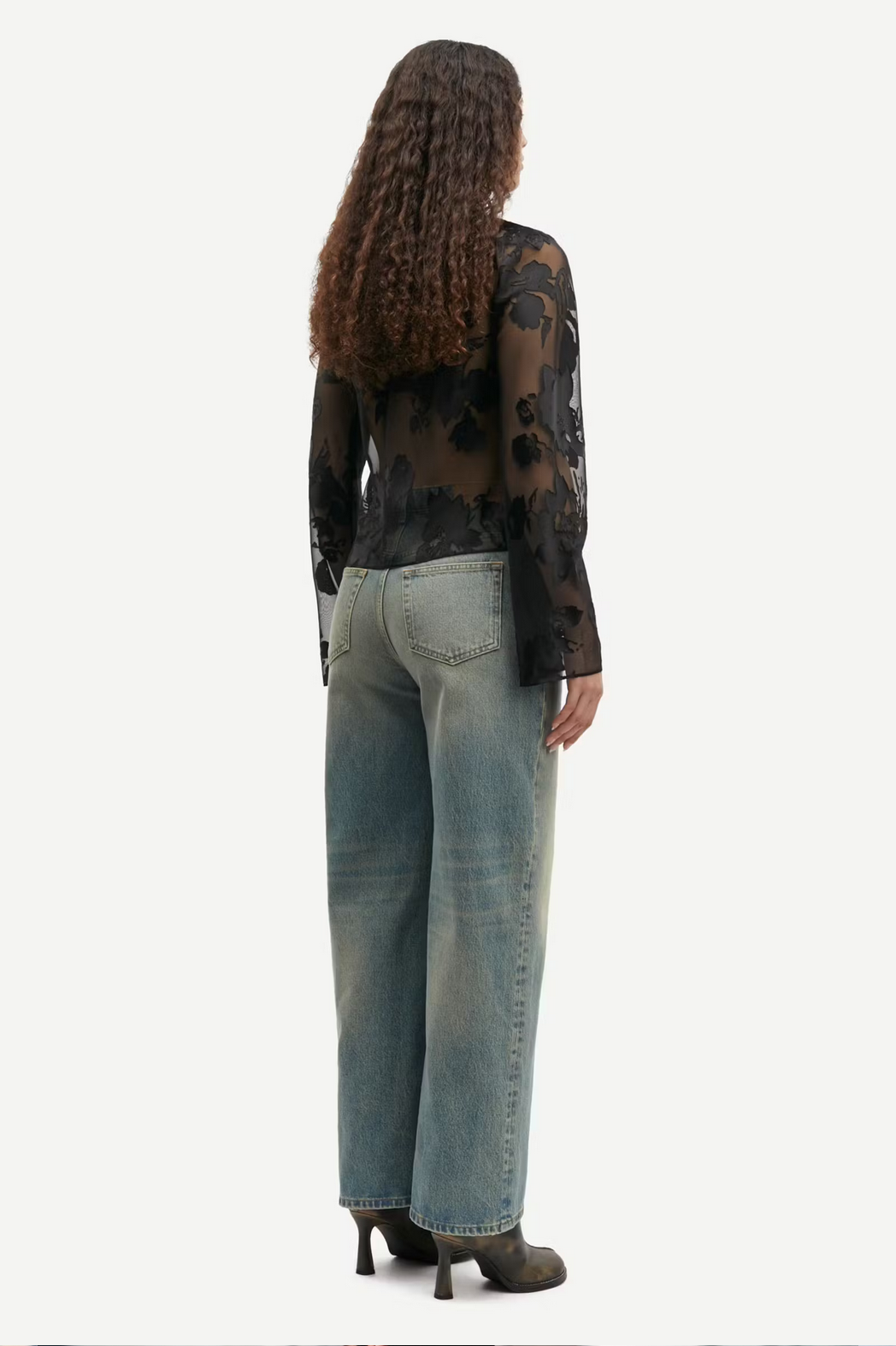 a women with brown hair wears a black sheer blouse with flower pattern and blue jeans samsoe samsoe