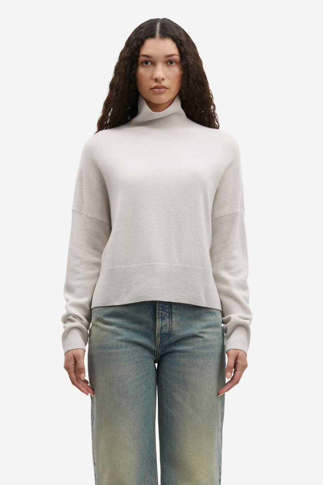 A person with long curly hair is wearing the Samsøe Samsøe Nola Cashmere Turtleneck in Turtledove and blue jeans, standing against a plain white background.