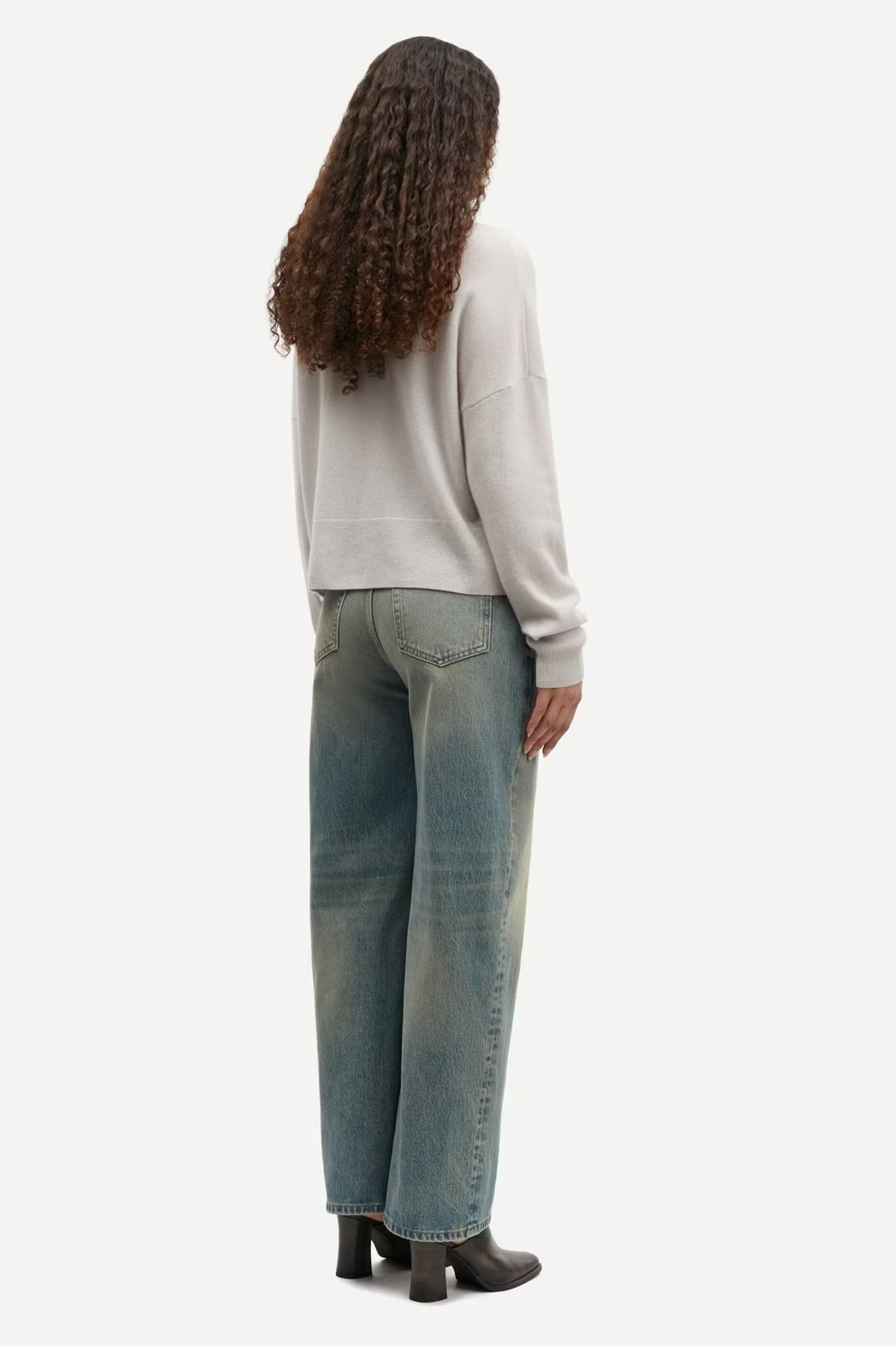 A person with curly hair wearing a Turtledove Nola Cashmere Turtleneck from Samsøe Samsøe, faded blue jeans, and black ankle boots stands facing away from the camera.