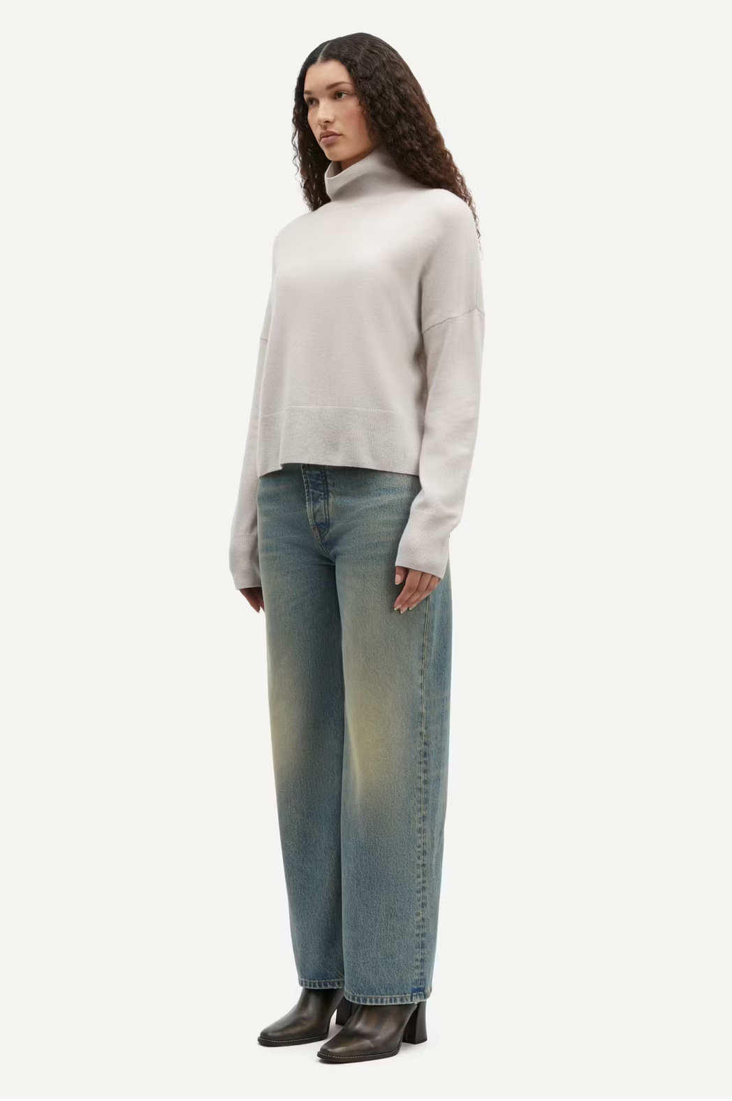 A person with long curly hair stands wearing a light-colored Nola Cashmere Turtleneck in the Turtledove shade from Samsøe Samsøe, paired with faded blue jeans and black shoes, facing slightly to the side against a plain white background.