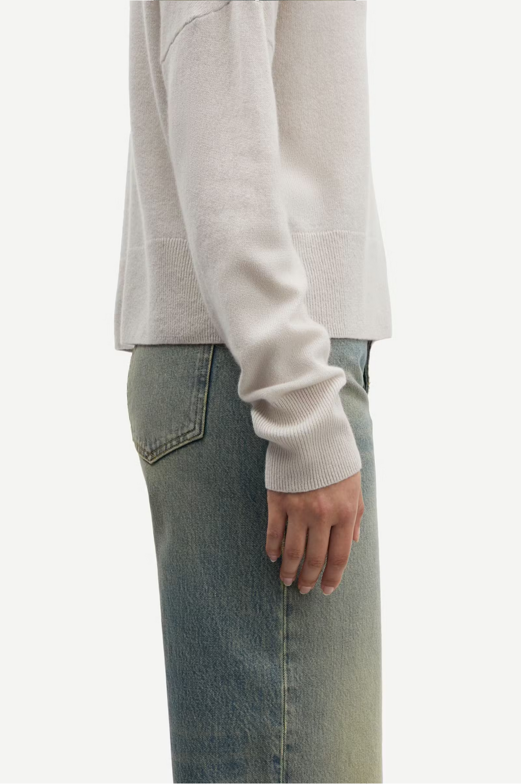 A person wearing a Samsøe Samsøe Nola Cashmere Turtleneck in Turtledove and faded blue jeans stands turned to the side, with their arm relaxed by their side.