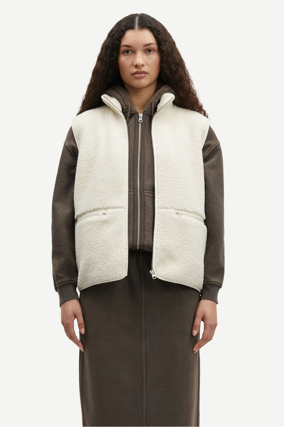 a woman with brown hair stands in a brown hoodie, skirt, and cream vest jacket by samsoe samsoe