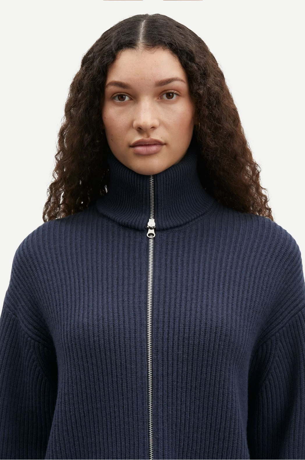 A person with curly hair is wearing the Samsøe Samsøe Nova Cardigan in an Outer Space shade, featuring a ribbed turtleneck design made of 100% wool and a sleek two-way zipper closure, standing against a plain background.