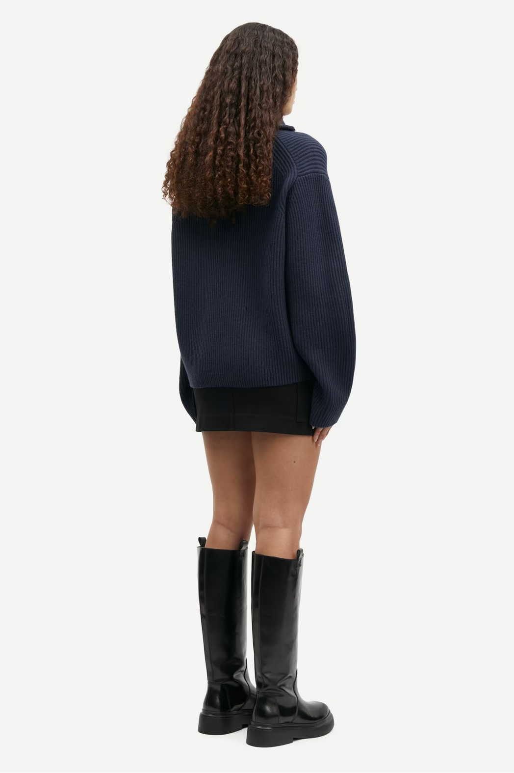 A person with long curly hair is wearing a dark blue Nova Cardigan - Outer Space by Samsøe Samsøe made from 100% wool, paired with a black skirt and black knee-high boots, facing away against a plain white background.