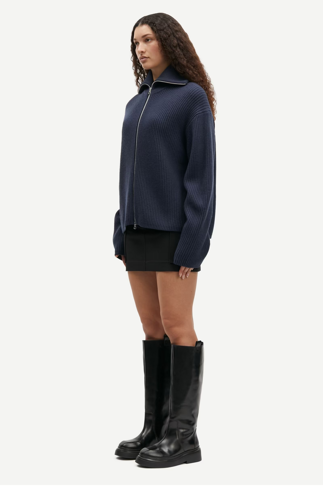 A person with curly hair is wearing the Samsøe Samsøe Nova Cardigan in Outer Space, which features a two-way zipper closure, paired with a black skirt and knee-high black boots, standing against a plain white background.