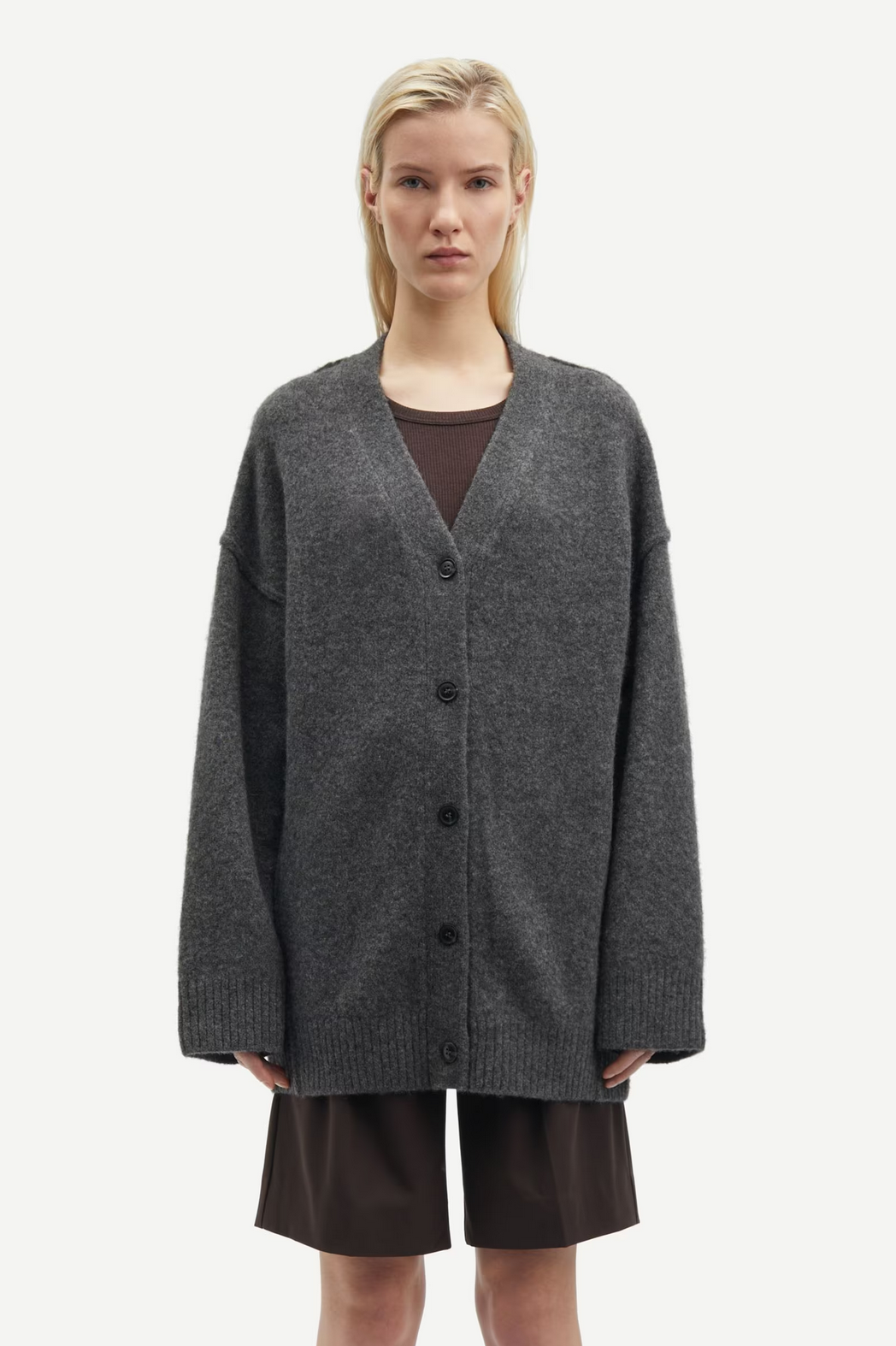 A person is wearing the Saceleste Cardigan in Black Oyster by Samsøe Samsøe, featuring a dark gray wool blend with dropped shoulders and black buttons, paired with brown shorts and standing against a plain white background.