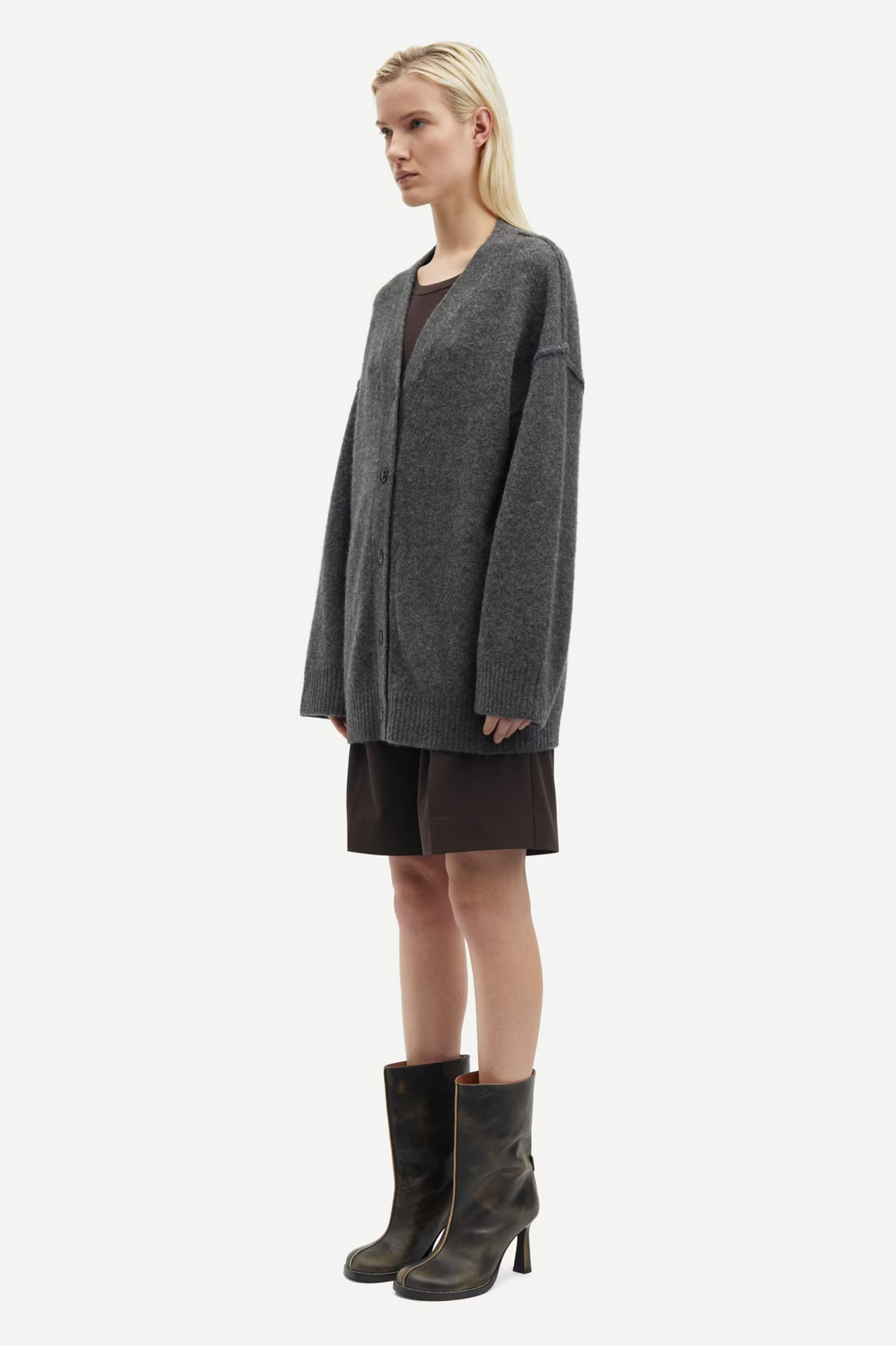 A person in a black oyster, oversized Saceleste Cardigan by Samsøe Samsøe with dropped shoulders and dark boots stands against a plain white background.