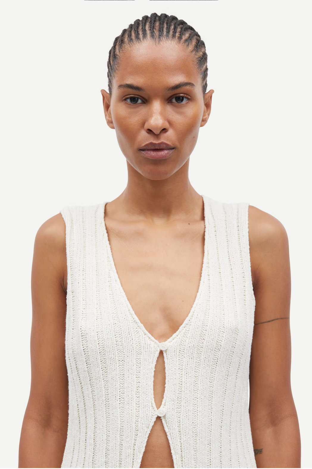 A model showcases the Salou Knit Vest - Egret by Samsøe Samsøe, a chic, sleeveless white V-neck top with a front opening. The plain white backdrop highlights its elegance and simplicity, making it ideal for any capsule wardrobe. Crafted from organic cotton.