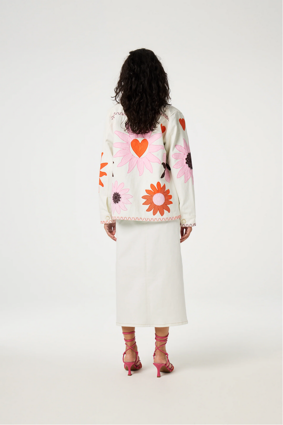 Elody, with her curly hair cascading down her back, stands gracefully on a plain background. She dons the Fabienne Chapot Elody Jacket in Warm White/Floral, paired with vibrant pink heels.