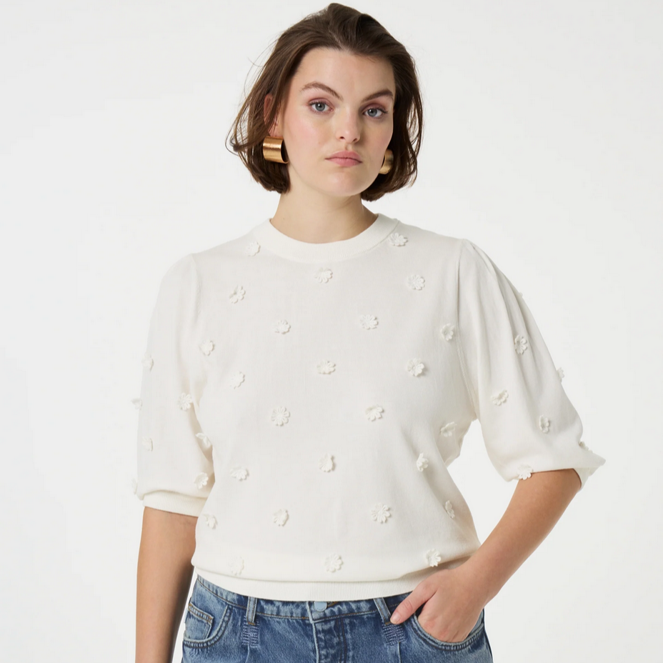 a woman stands and wears a white sweater with floral applique details by fabienne chapot and blue jeans on a back ground