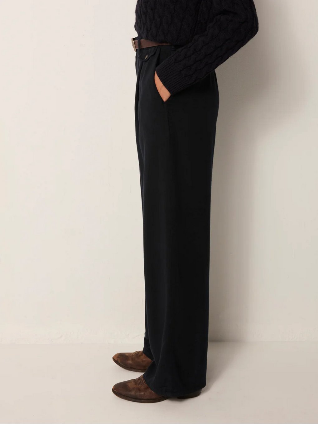 A person standing sideways wears Maison Hotel's Keaton Trousers in Night Blue, styled with a dark textured sweater, a brown belt, and brown pointed shoes.