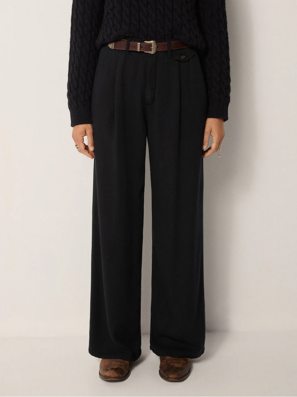 Person wearing Maison Hotel’s Keaton Trousers in Night Blue, paired with a cable-knit sweater, brown belt, and brown shoes, standing against a plain light background.