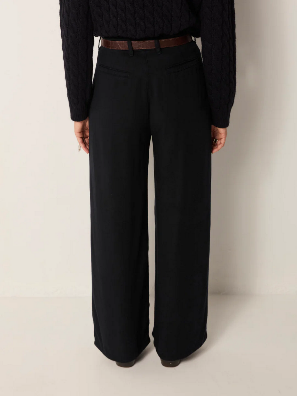 A person is shown from the back against a plain background, wearing Maison Hotel's Keaton Trousers in Night Blue, paired with a dark cable knit sweater and a brown belt.